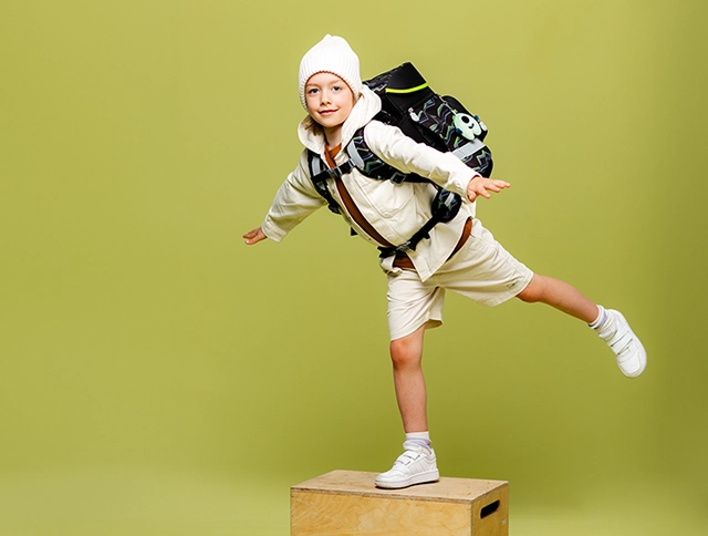 Kind mit Schulranzen auf dem Rücken balanciert mit einem Bein auf einem Kasten