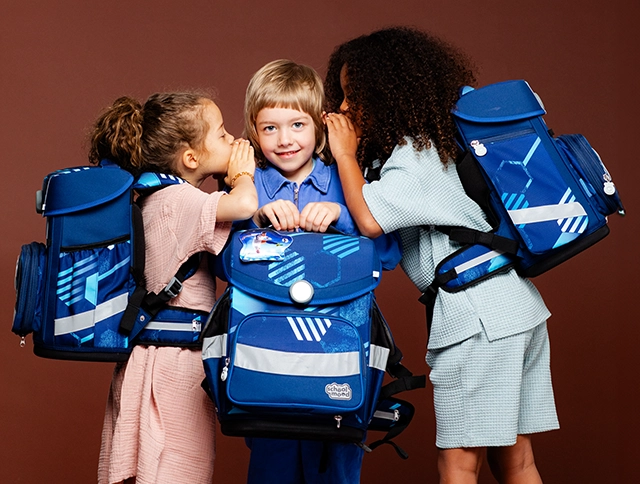 Kind in der Mitte mit Schulranzen in der Hand wird von zwei Kindern mit Schulranzen auf dem Rücken in die Ohren geflüstert
