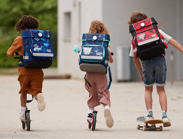 Zwei Kinder auf Cityroller und ein Kind auf Skateboard mit Schulranzen auf dem Rücken von hinten