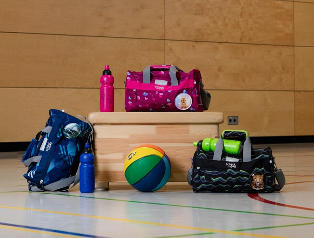 Sporttaschen und Flaschen rund um und auf einem Kasten in einer Turnhalle
