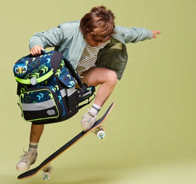 Kind mit Schulranzen in der Hand macht einen Skateboard-Trick
